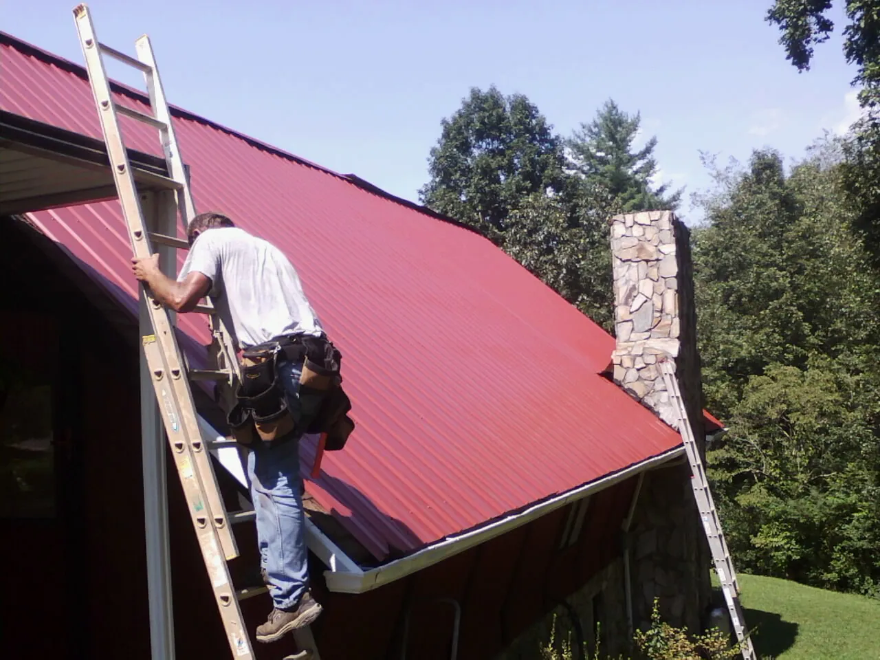 Roofer on a ladder beside a red metal roof and stone chimney — Soffit & Fascia Repair by Roofing Brothers in Cohutta